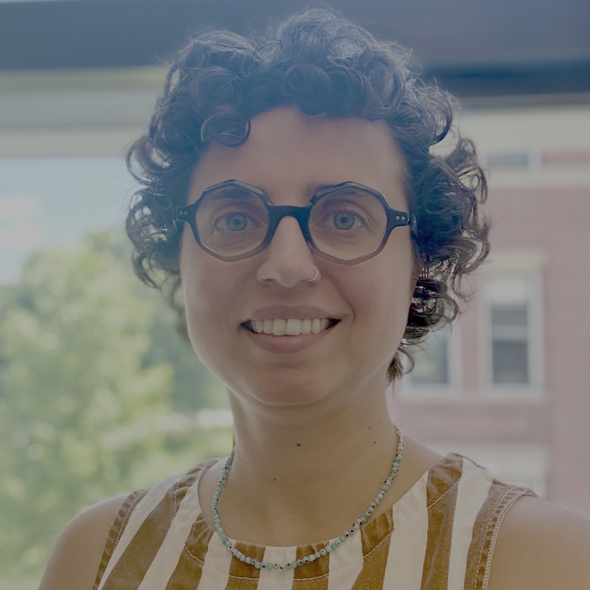 A headshot of a woman with curly, short hair with green eyes and small almost-round glasses.